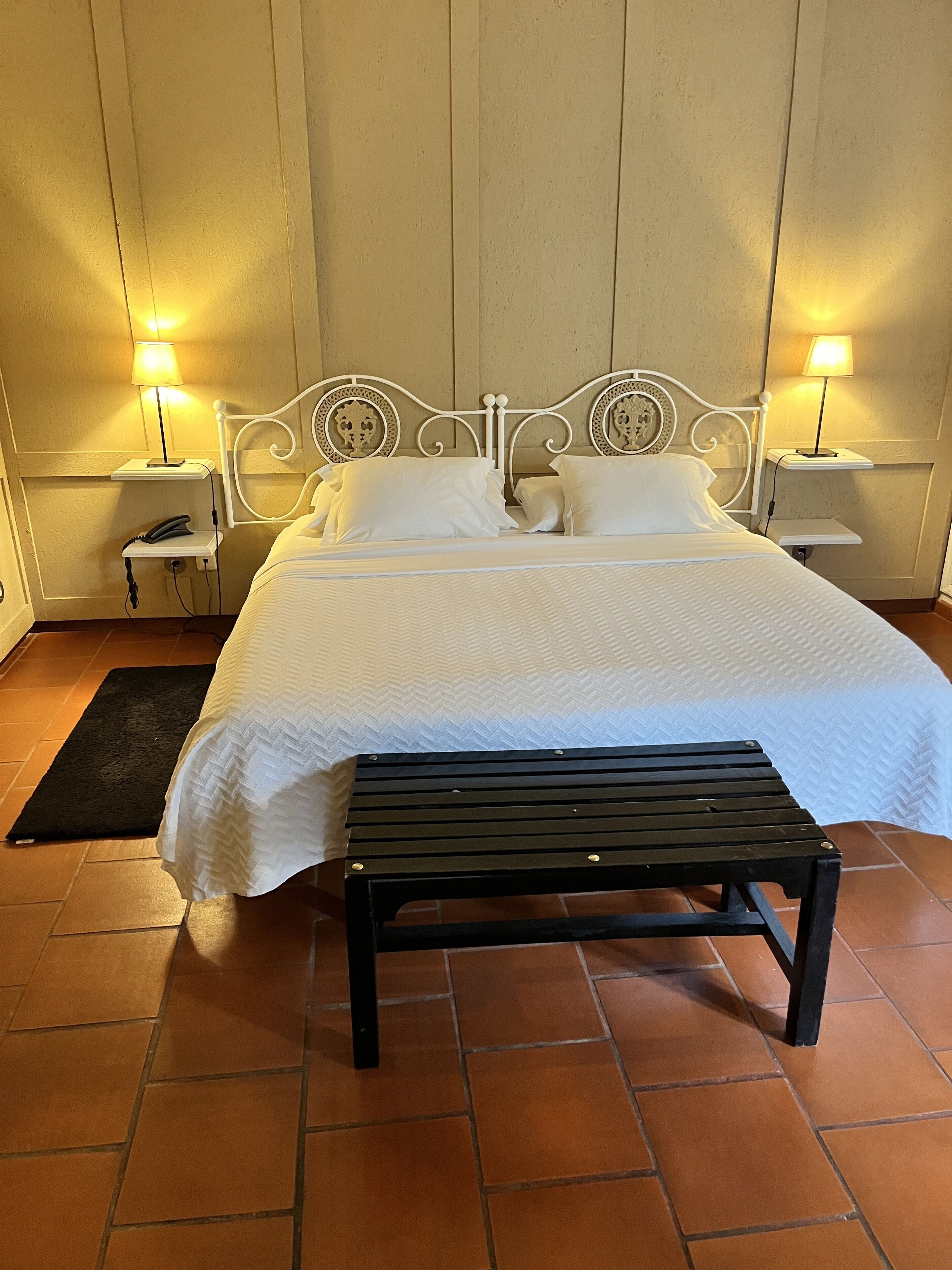A neatly made bed with white linens, a black bench, and two bedside lamps on matching nightstands.