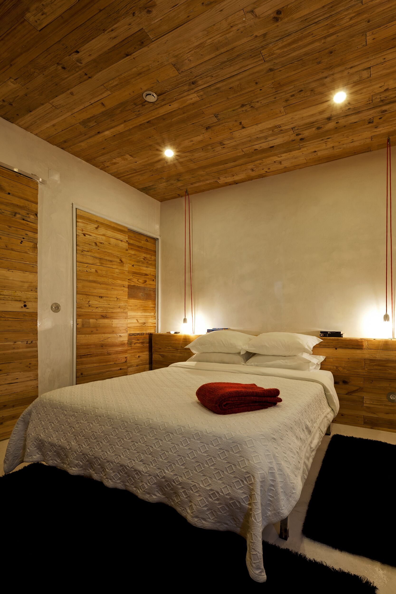 Un dormitorio con techo de madera, una cama con ropa de cama blanca, un cojín rojo y una alfombra negra.