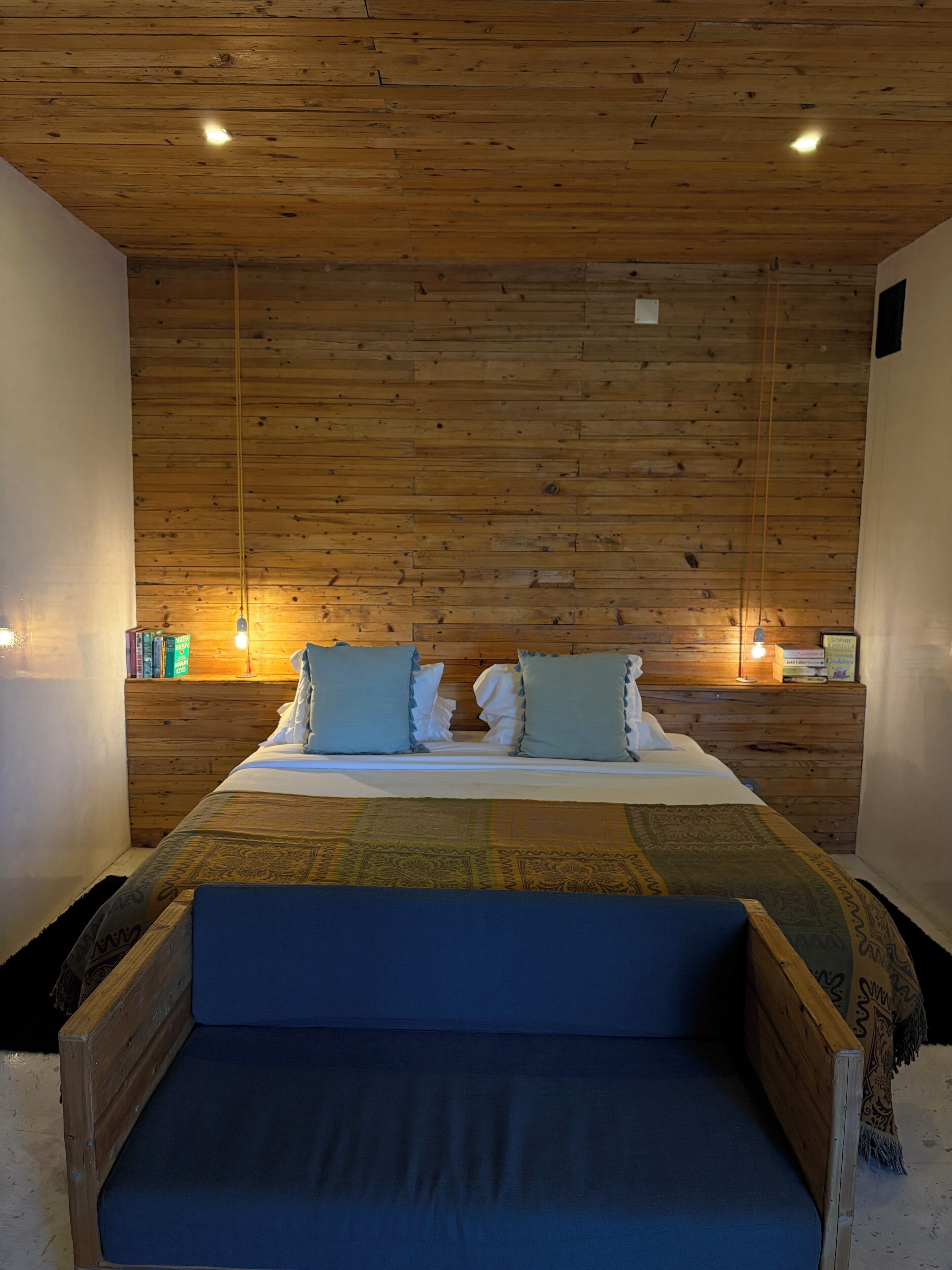 Un dormitorio con techo de madera, una cama con ropa de cama azul y dos lámparas de noche.