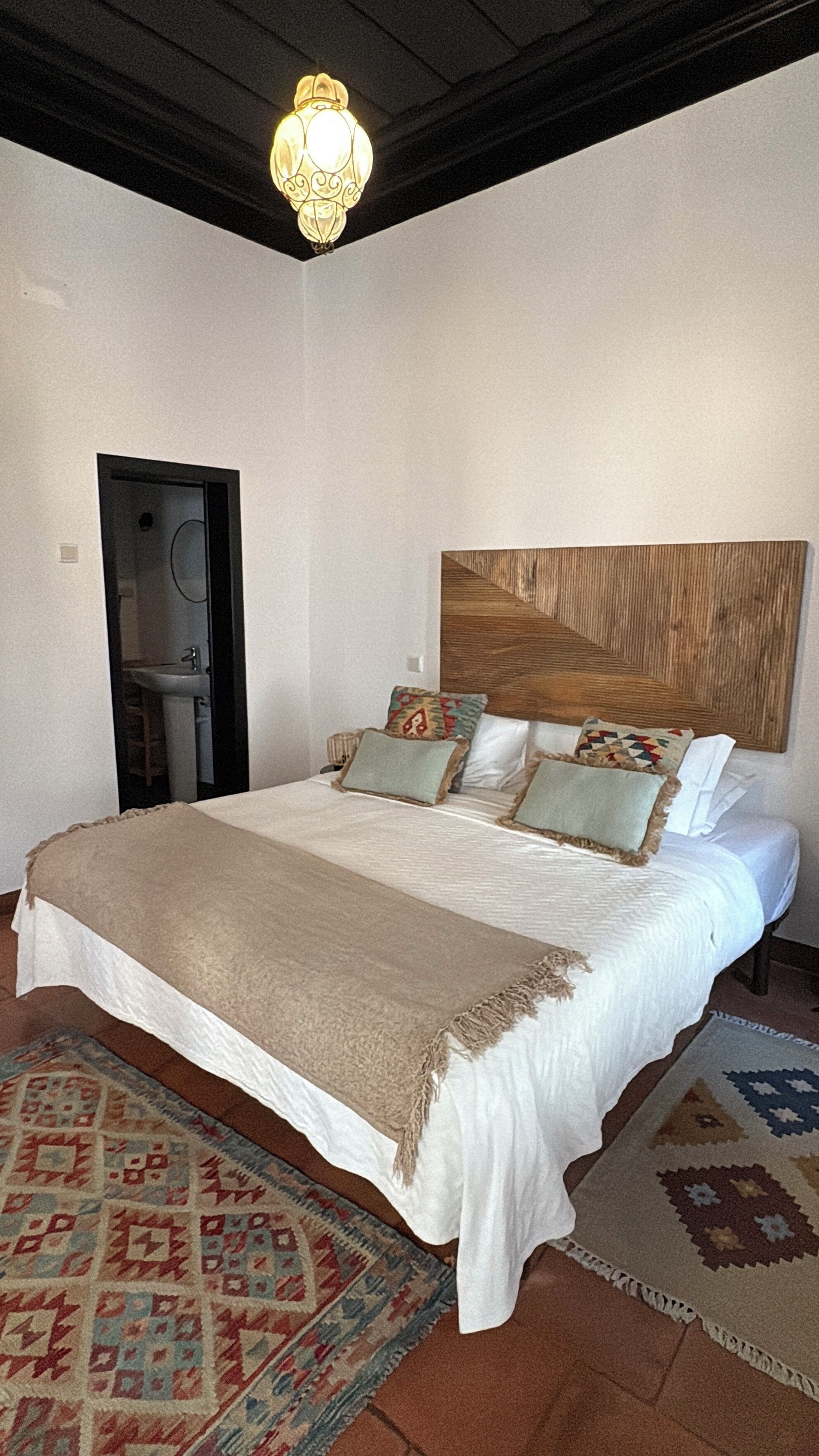 A bedroom with a large bed, a wooden headboard, a bedside table, a nightstand, a floor lamp, a patterned rug, and a door leading to a bathroom.