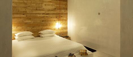 A bedroom with a wooden ceiling, a bed with white bedding, and a small shelf with books.