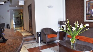 Ein Hotel-Empfangsbereich mit einer gebogenen Marmortheke, einem grauen Sessel mit orangefarbenem Kissen, einer Vase mit weißen Blumen und einem gerahmten Kunstwerk an der Wand.