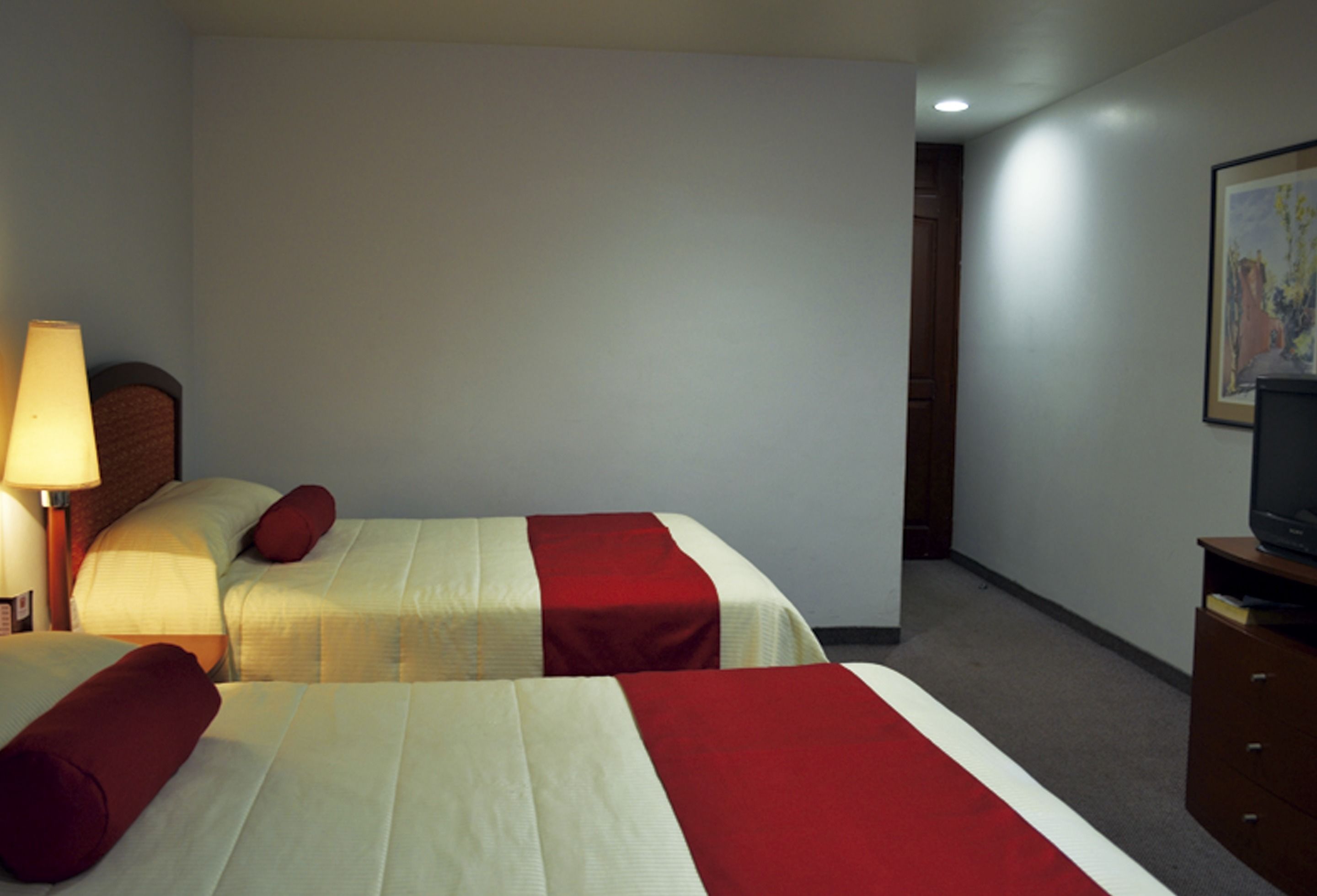 Een hotelkamer met twee bedden, een lamp, een televisie op een houten kast en een schilderij aan de muur.
