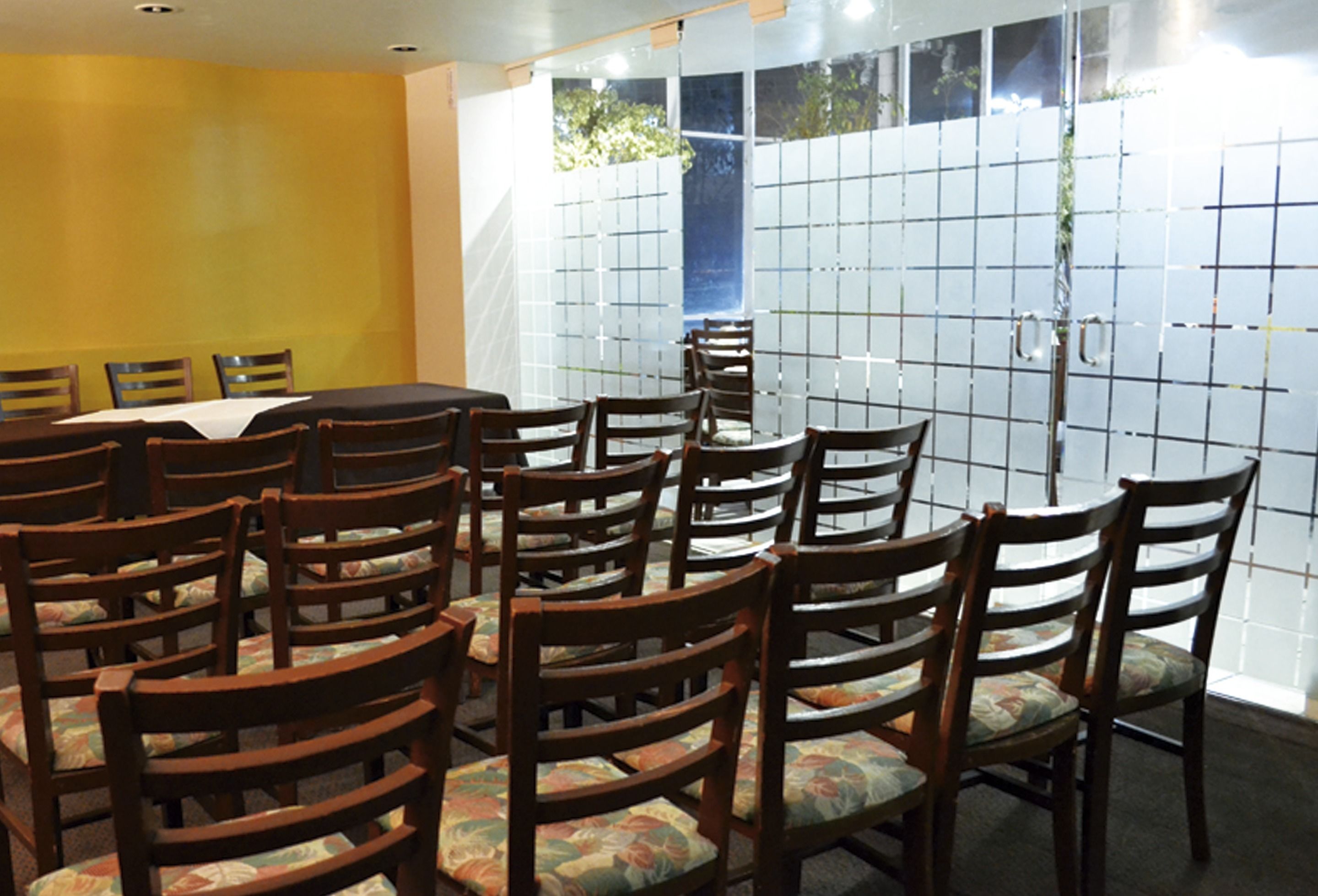 Un salón de conferencias con una gran mesa y varias sillas, una pared amarilla y un ventanal con vistas a árboles.