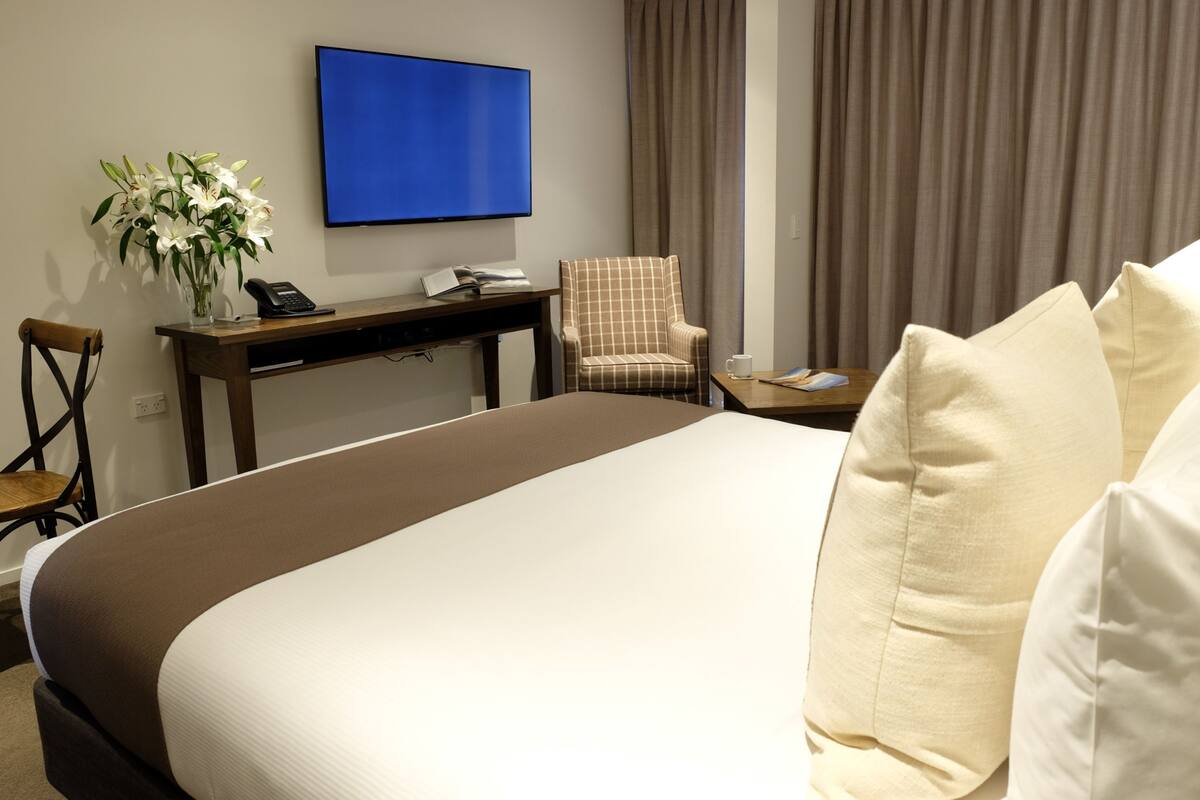 A hotel room with a large bed, a desk with a telephone, a chair, a TV mounted on the wall, and a window with curtains.