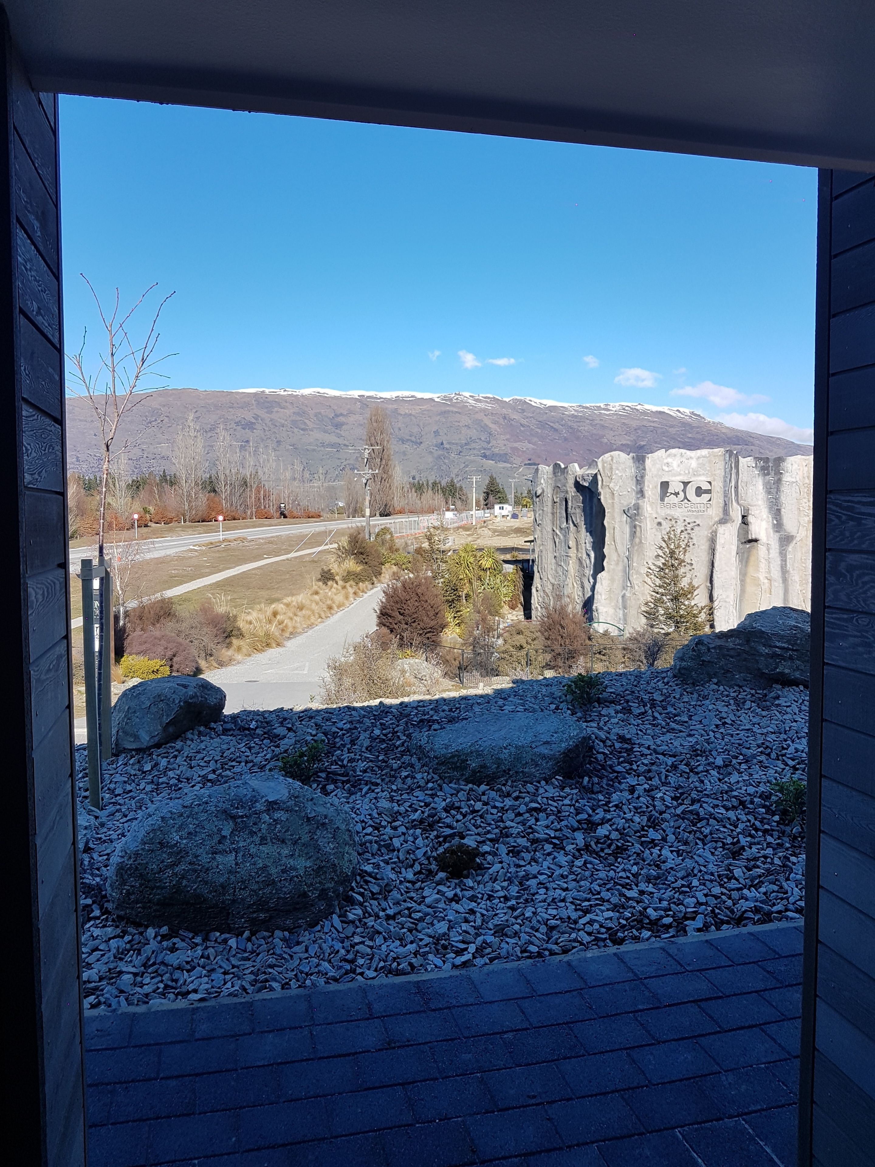 A view from a window overlooking a gravel area with a paved path, a stone wall, and a road leading to a mountain range with snow-capped peaks.