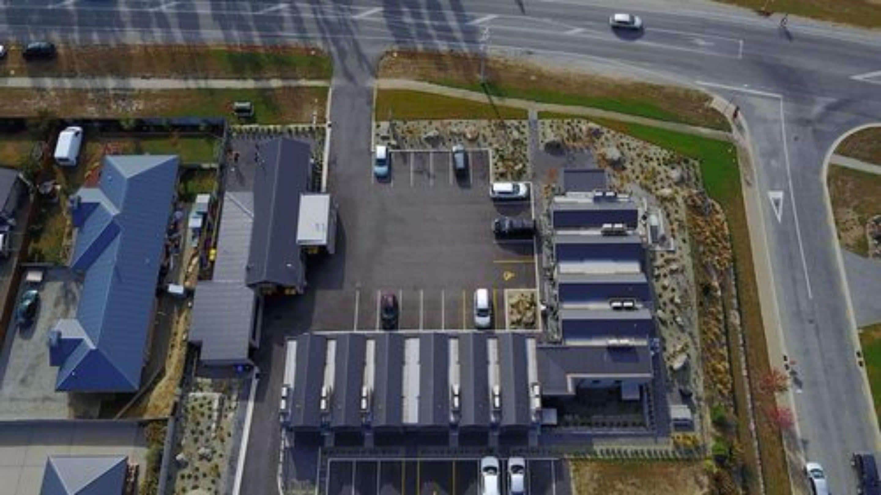 Une vue aérienne d’un parking avec plusieurs rangées de voitures garées, d’un bâtiment doté d’un toit plat et d’une route qui tourne vers la droite.