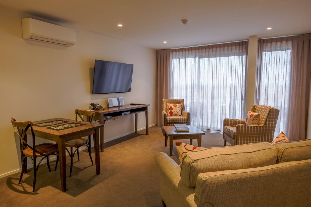 A hotel room with a flat-screen TV mounted on the wall, a dining table with chairs, a sofa, and a small coffee table.