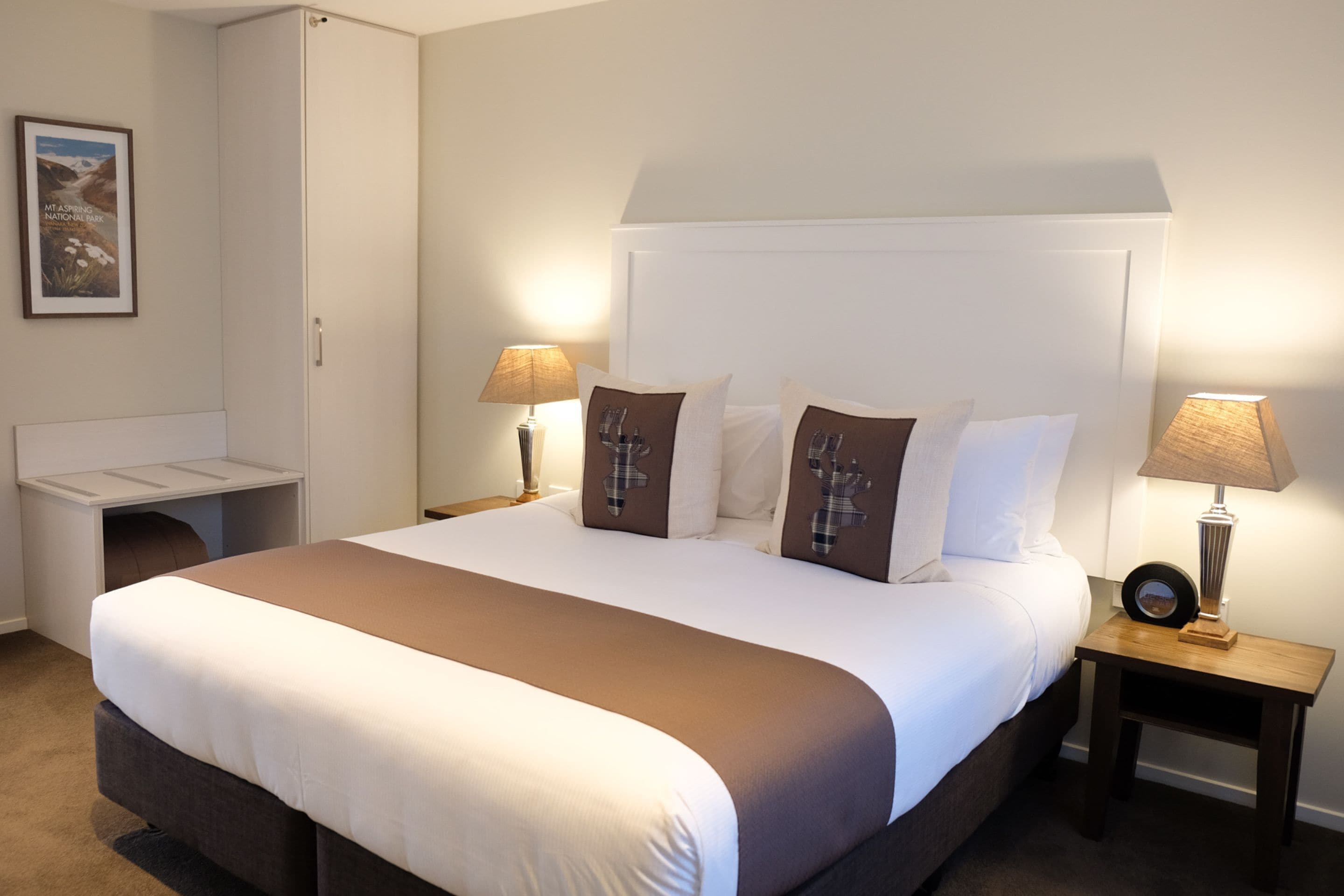 A neatly made bed with a headboard, two bedside tables with lamps, a small seating area, and a framed picture on the wall.