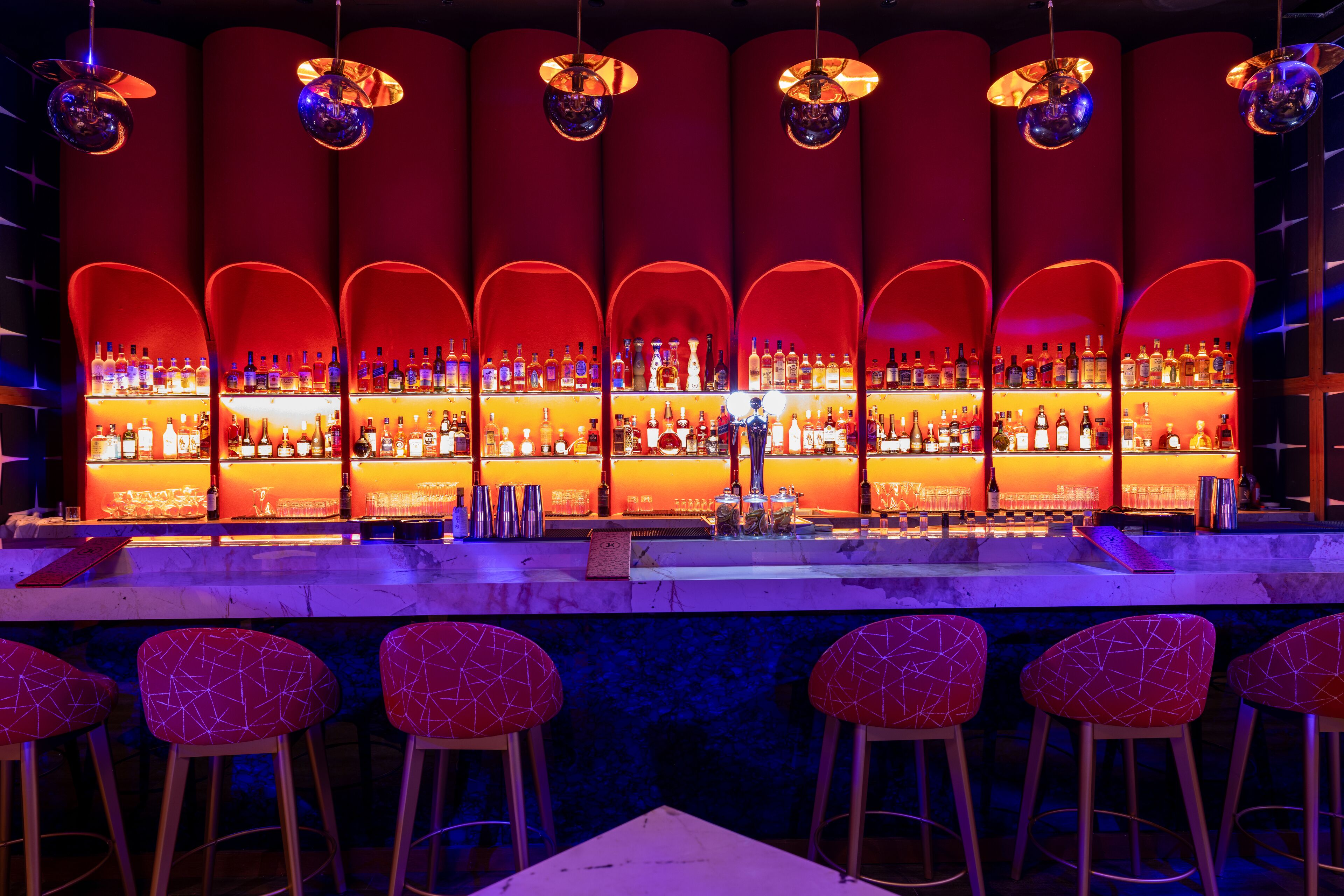 Un bar con varias estanterías llenas de botellas, taburetes rojos y luces de discoteca.