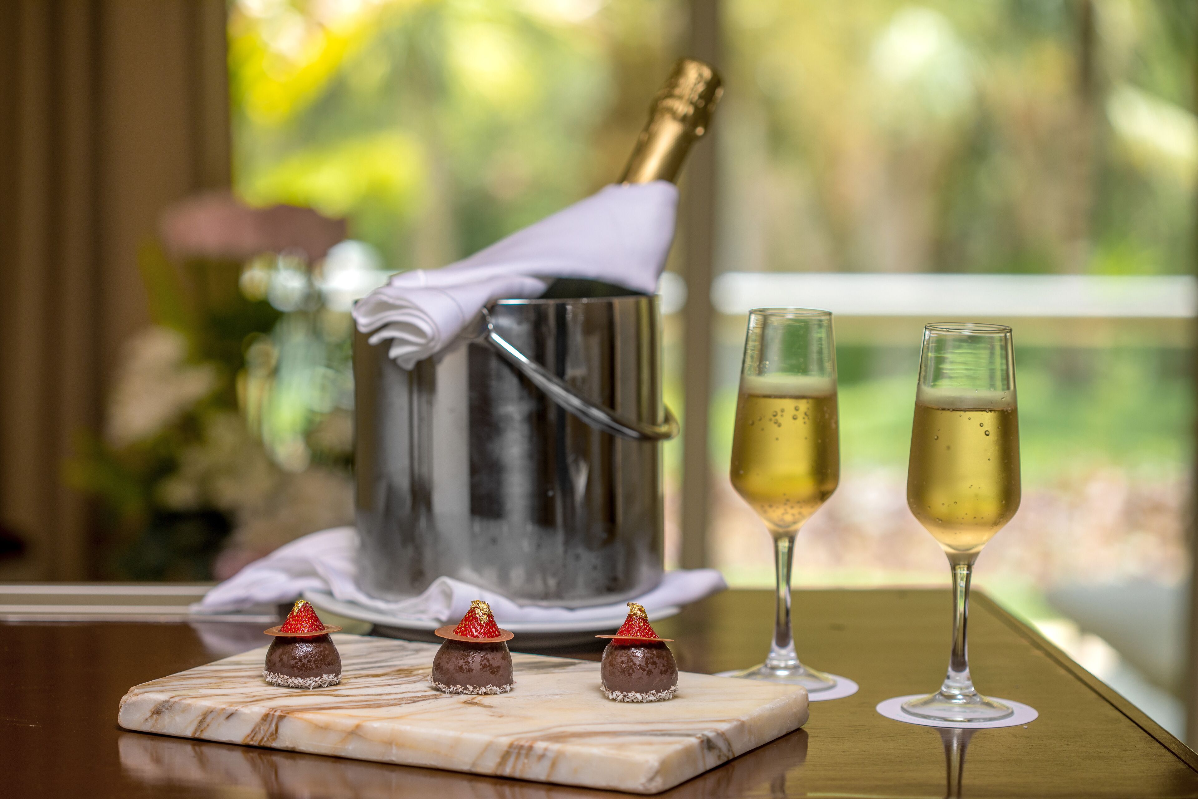 A table with two glasses of champagne, a bottle of champagne, and three chocolate-covered strawberries on a marble board.