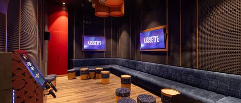 Een moderne lounge met een blauwe bank, houten stoelen en twee schermen waarop het woord 'KASSETTE' staat.