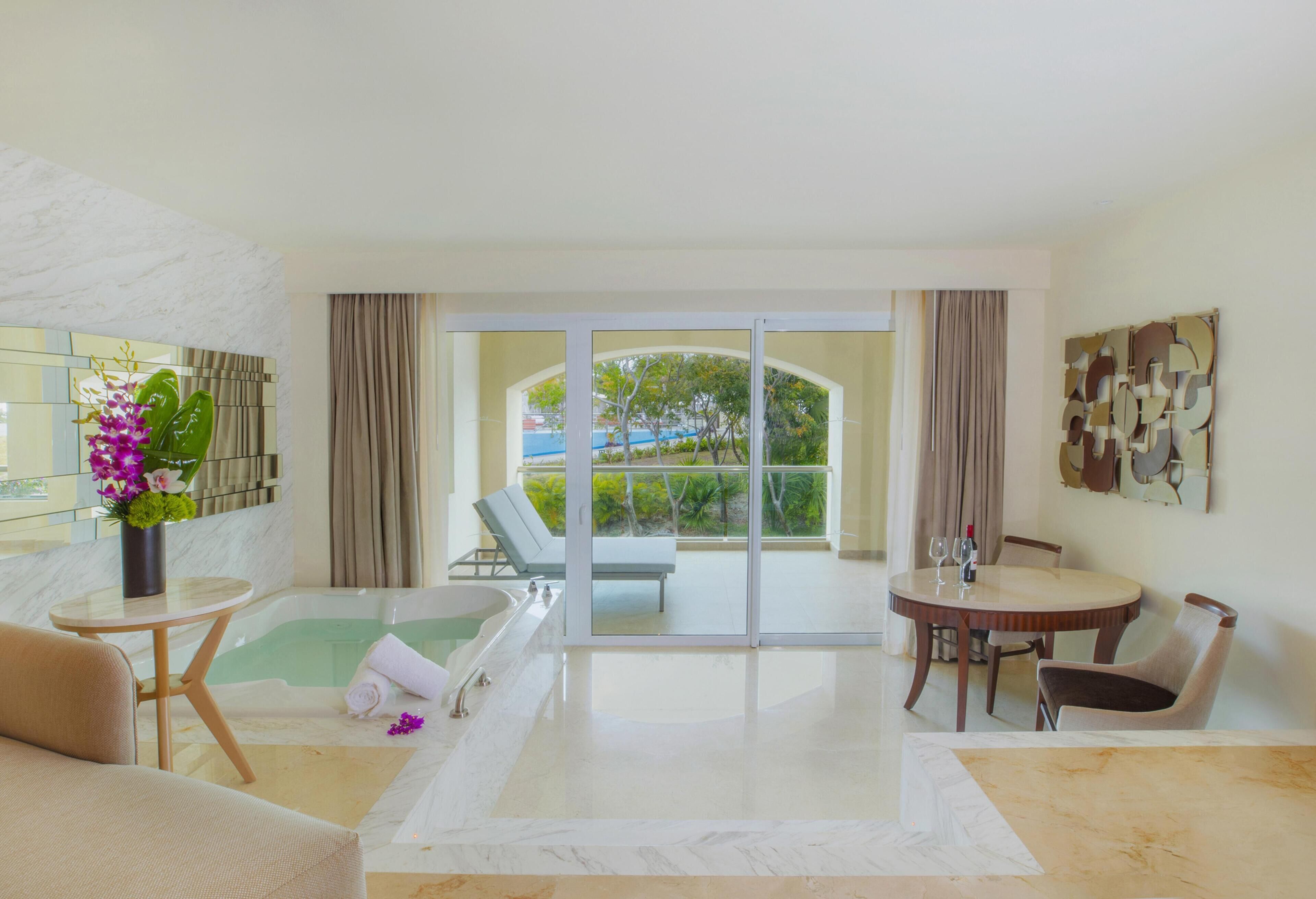 A modern hotel room with a glass door leading to a balcony, a bathtub, a dining table, and a sofa.