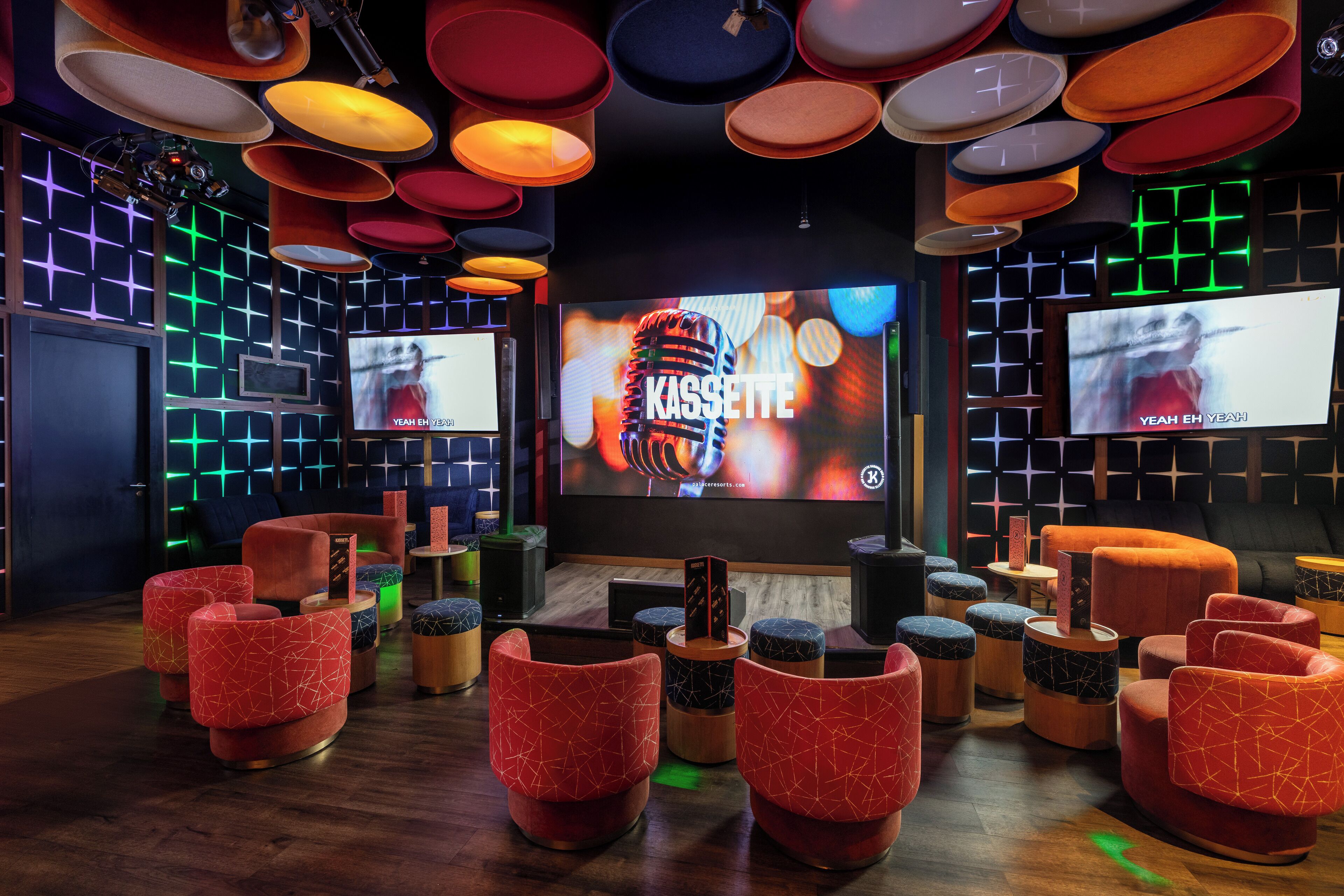 Un moderno espacio lounge con varias opciones de asientos, incluyendo sillas con patrones geométricos en rojo y azul, y una pantalla grande que muestra la palabra "KASSETTE".