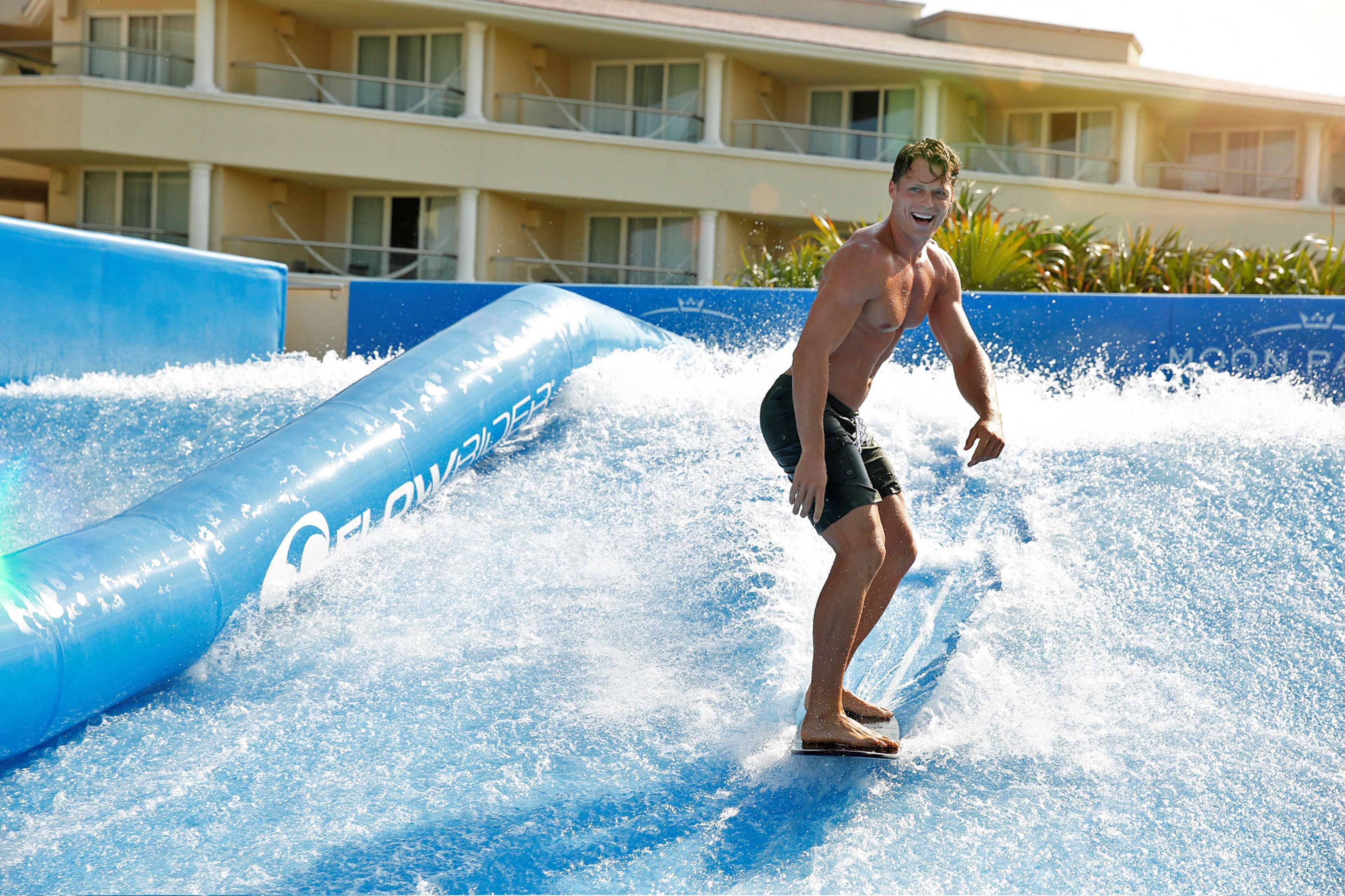 A person surfing on a wave in a water park area with a building in the background.