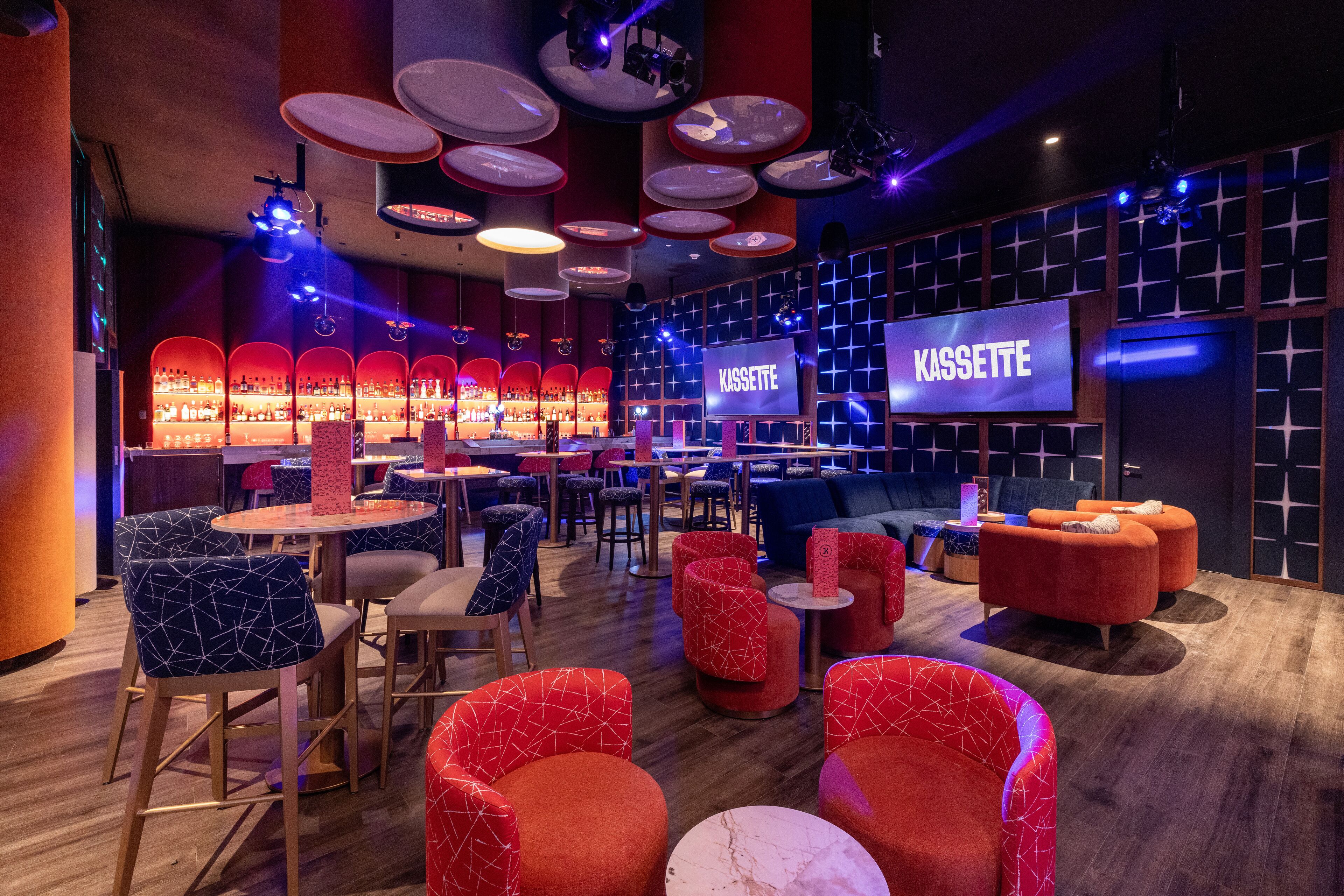 Un espace bar moderne avec des sièges aux motifs rouges et bleus, un panneau indiquant « KASSETTE » et un comptoir bien garni de bouteilles.