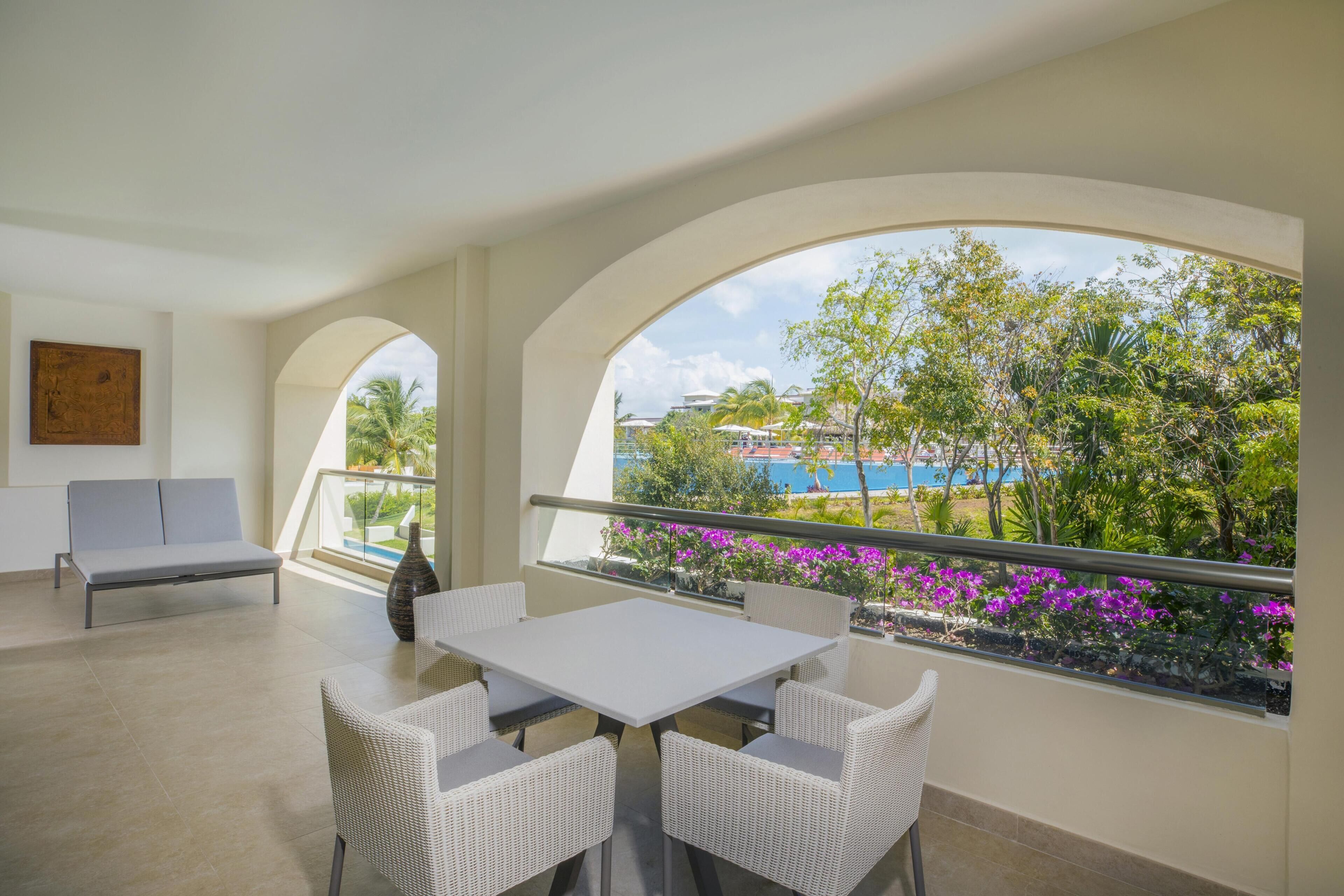 A balcony with a white table and chairs, a sofa, and a view of a pool and greenery.