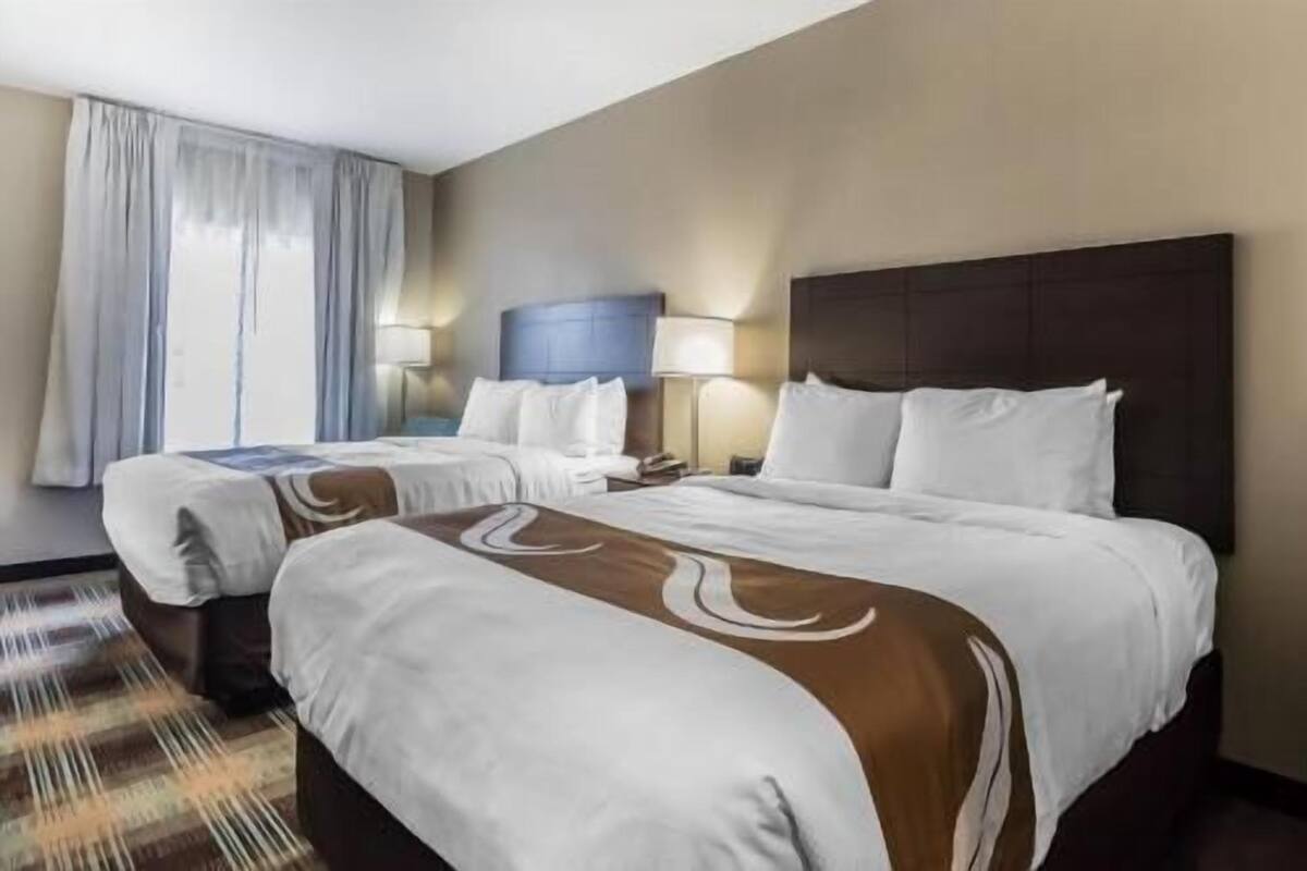 Ein Hotelzimmer mit zwei Betten, einem großen Fenster mit Vorhängen und einer Kopfteil.