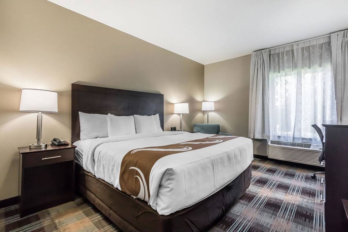 Ein Hotelzimmer mit einem großen Bett, zwei Nachttischlampen, einem Schreibtisch und einem Fenster mit Vorhängen.