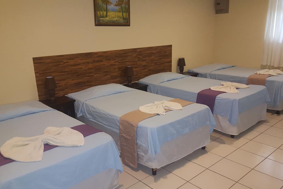 A hotel room with four single beds, each with a blue bedspread and a white towel folded on top. The room has a wooden headboard, a painting on the wall, and a window with curtains.