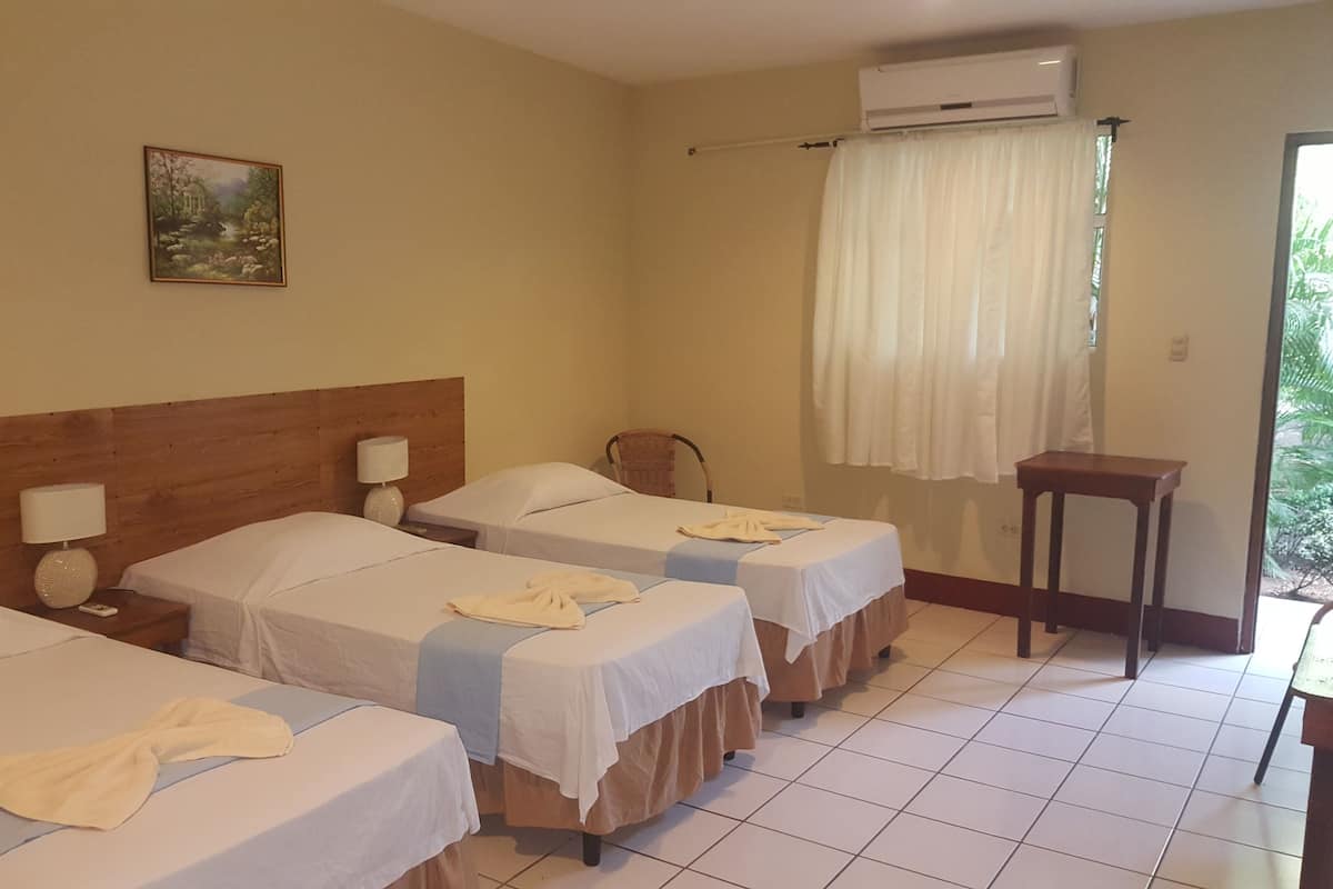 A hotel room with three single beds, a wooden headboard, a painting on the wall, an air conditioning unit, and a window with curtains.