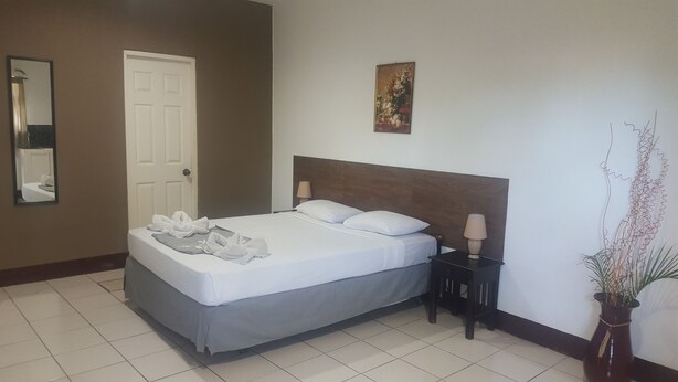 Habitación de hotel con cama, mesita de noche, lámpara, espejo, puerta y un cuadro en la pared.