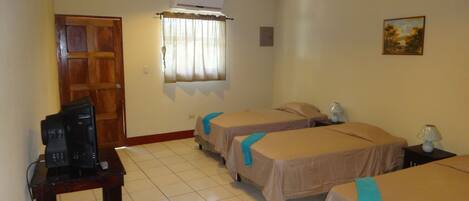 Habitación de hotel con dos camas, un ventilador de techo, un televisor sobre una mesa de madera y una ventana con cortinas.