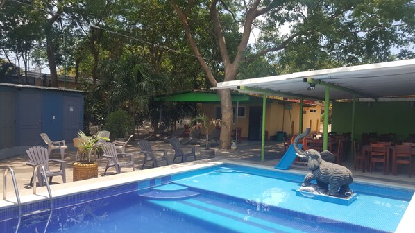 Una piscina con tobogán, rodeada de sillas de descanso y una zona de estar cubierta.