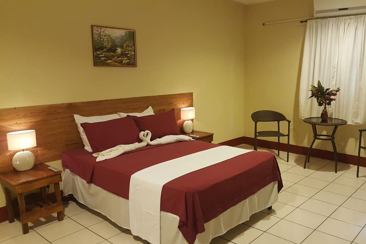 A hotel room with a bed, two bedside tables, a nightstand, a small round table with chairs, and a window with curtains.