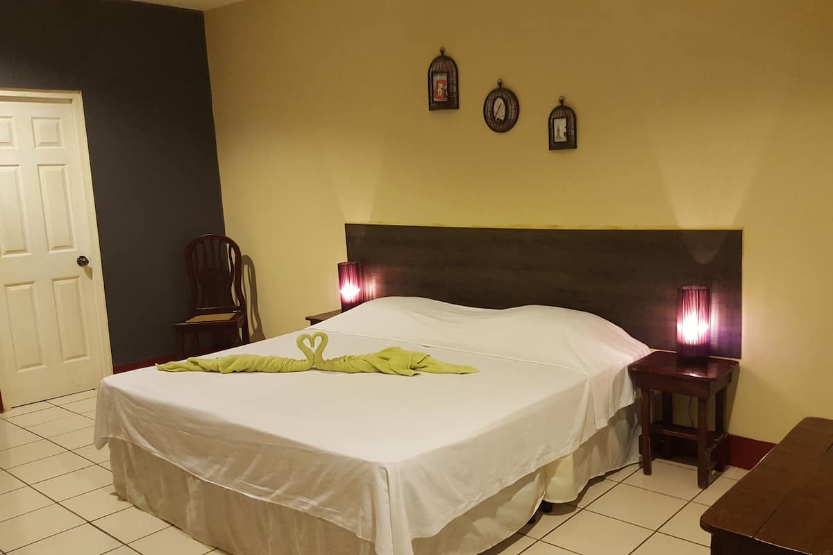 A neatly made bed with a white bedspread and a green towel shaped like a heart. There are two bedside lamps, a wooden nightstand, and a chair.