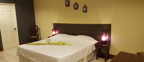 A neatly made bed with a white bedspread and a green towel shaped like a heart. There are two bedside lamps, a wooden nightstand, and a chair.