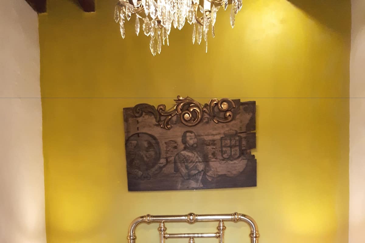 Een bed met metalen frame en beddengoed met een patroon, een houten hoofdeinde, een kroonluchter en twee bedlampjes.