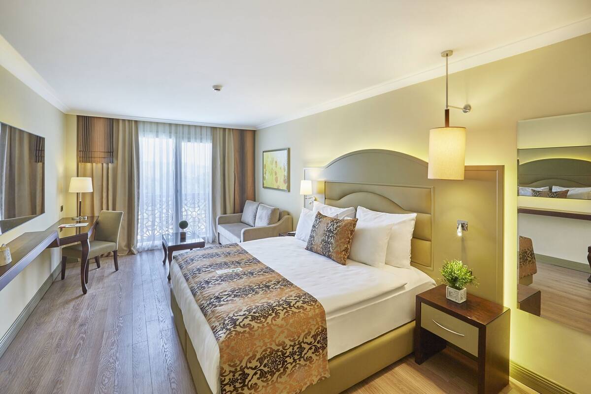A hotel room with a large bed, a desk, a chair, a bedside table with a lamp, and a mirror.