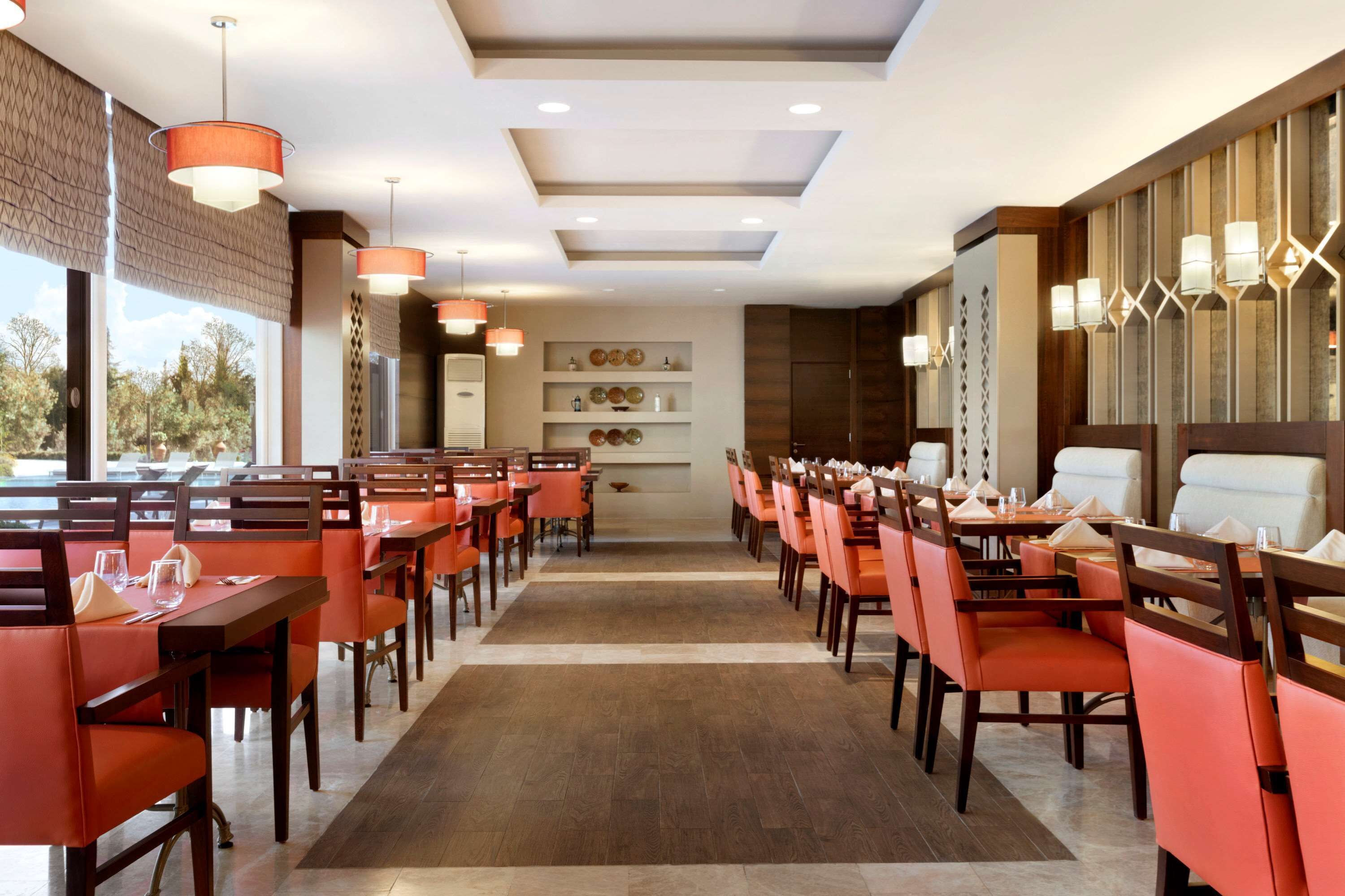 A restaurant with long tables and orange chairs, a wooden floor, and a view of trees outside.