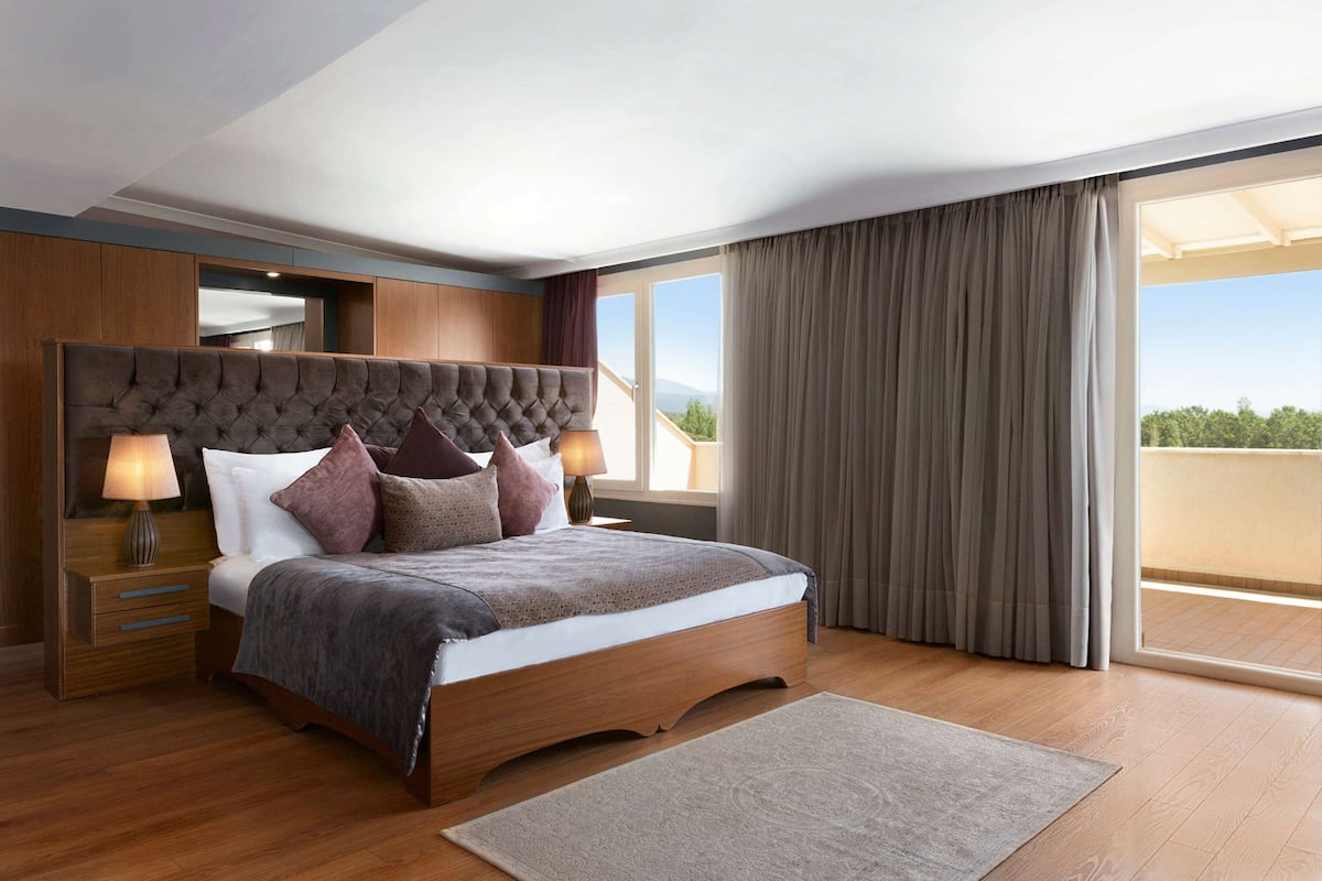 A modern bedroom with a large bed, wooden headboard, and a view of the outdoors through large windows.