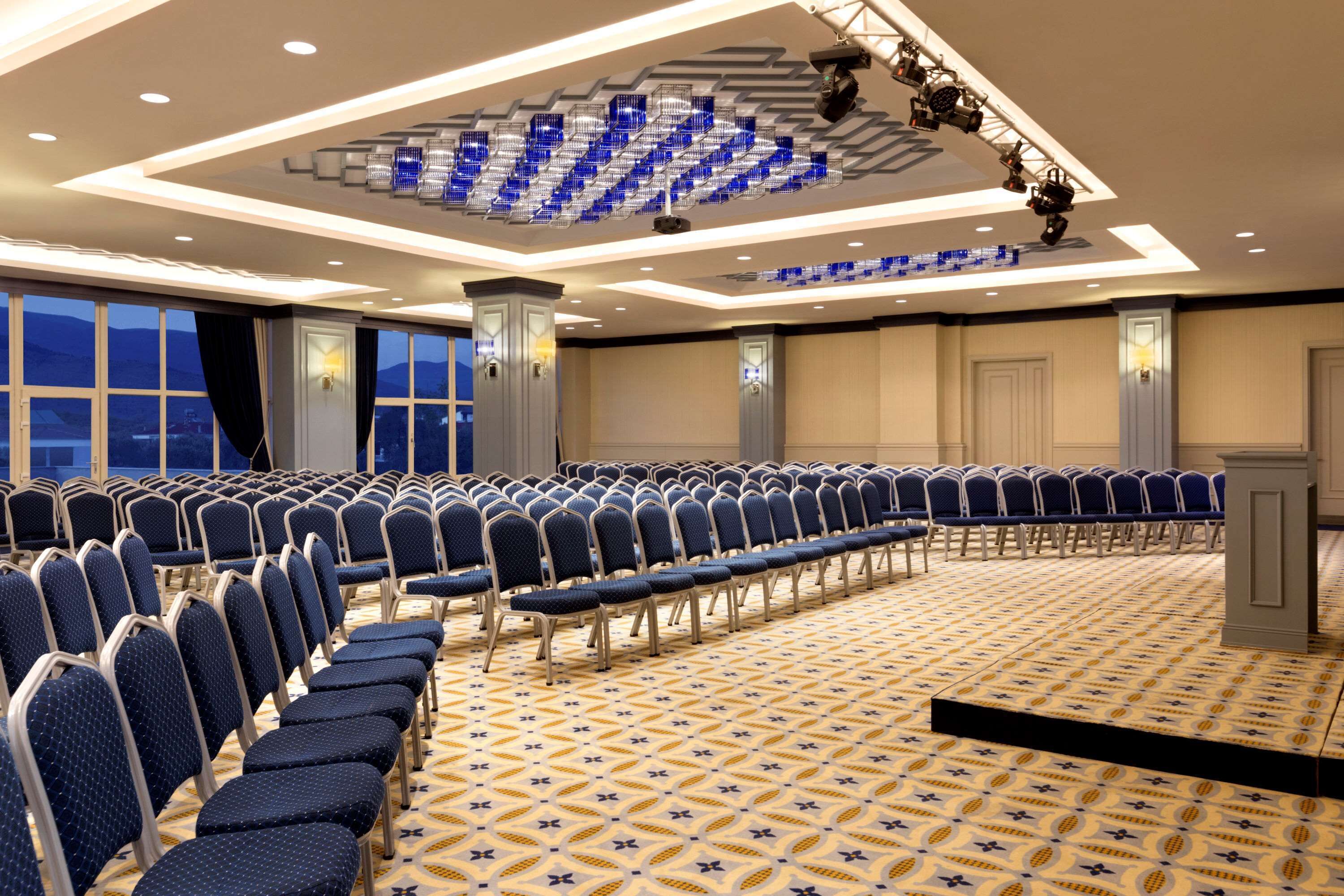 Un salón de conferencias con hileras de sillas, alfombra estampada y diseño de techo moderno.