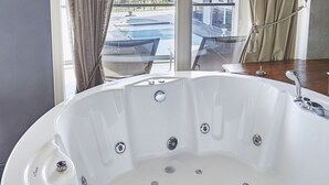 A modern bathroom with a large white bathtub, beige curtains, and a view of a swimming pool and buildings.