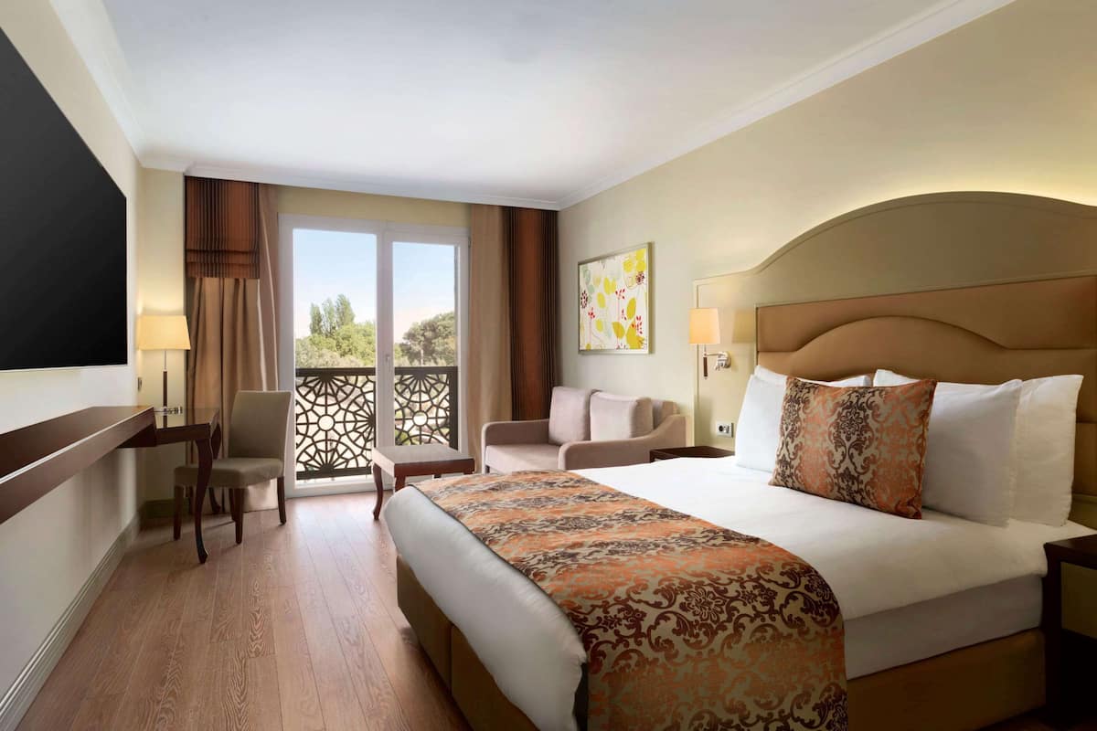 A hotel room with a large bed, a desk, a chair, and a balcony with a view.