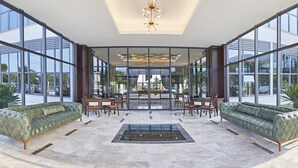 A spacious lobby with a high ceiling, large windows, a central chandelier, and a seating area with sofas and chairs.