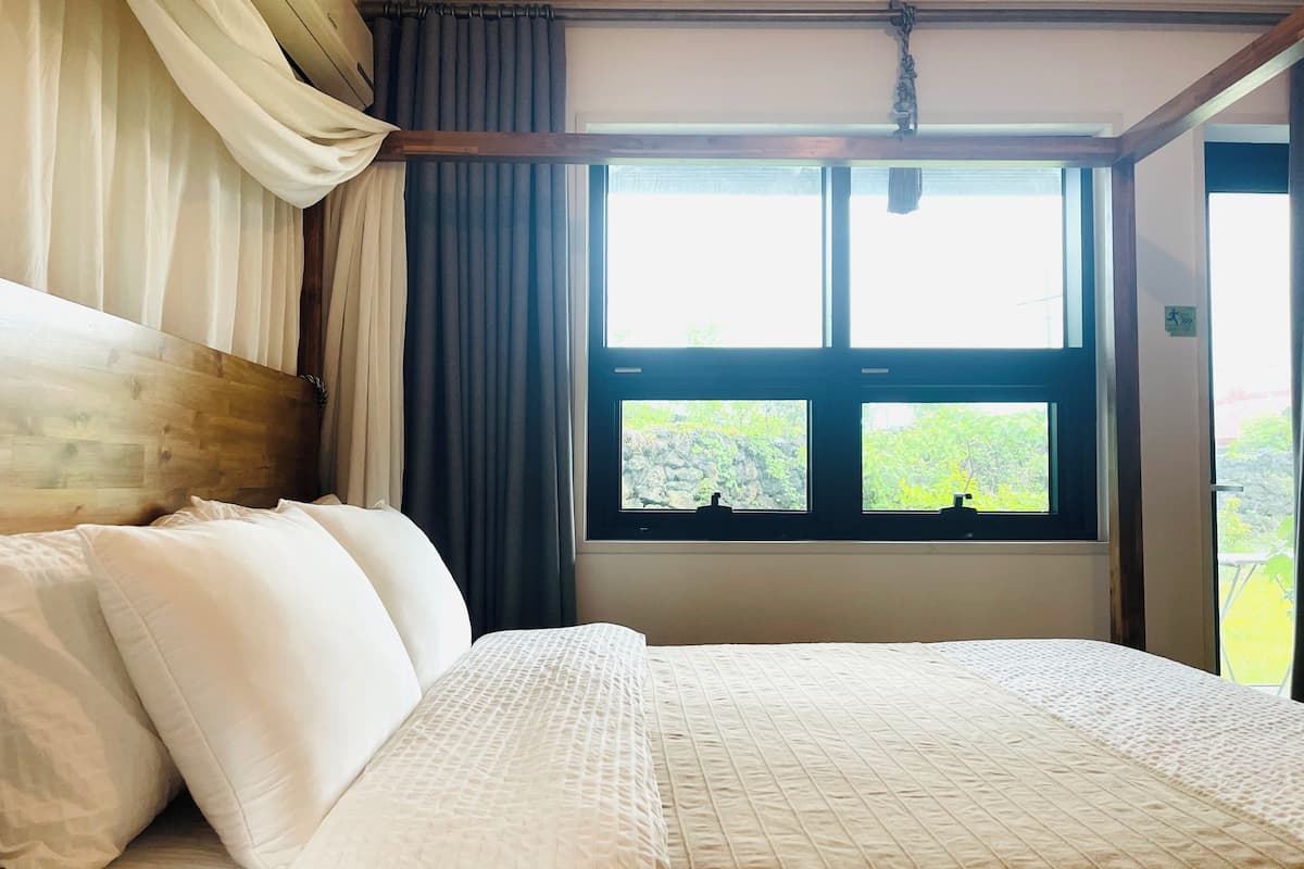 A hotel room with a large bed, a wooden headboard, a window with a view of greenery, and a curtain.
