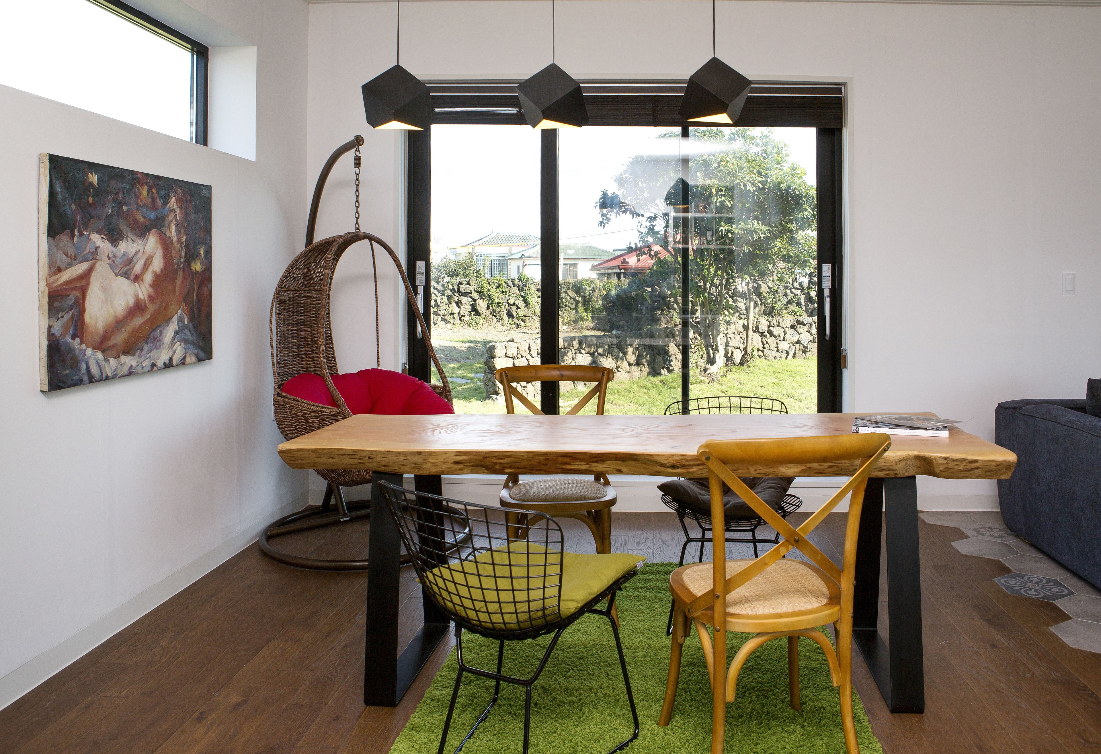 Un coin repas avec une table en bois, des chaises et un fauteuil suspendu. Un tableau orne le mur et de grandes fenêtres offrent une vue sur l’extérieur.