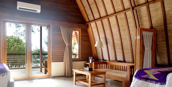 Ein Zimmer mit Holzbalkendecke, einem Bett, einem Tisch und einer Bank. Ein Fenster mit Vorhängen bietet Blick auf viel Grün draußen.