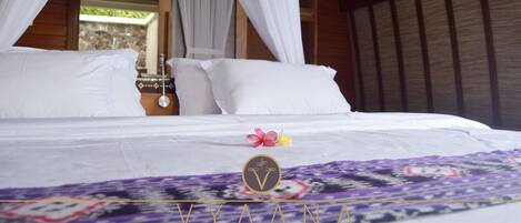 A bed with white linens, a purple and white patterned bedspread, and a pink flower on the bed.