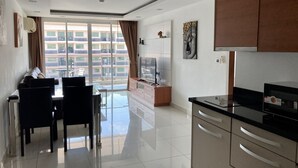 A modern kitchen with a black countertop and stainless steel appliances, a dining area with a glass table and chairs, and a large window with a view of a residential building. - Hyde Park Residence by PSR ASIA (Pattaya)