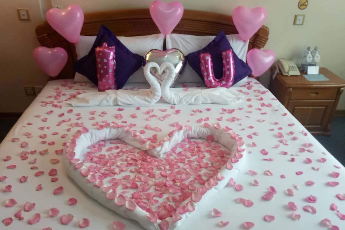 Ein Bett mit Rosenblättern und herzförmigen Dekorationen, inklusive Ballons mit der Aufschrift 'I LOVE YOU'.