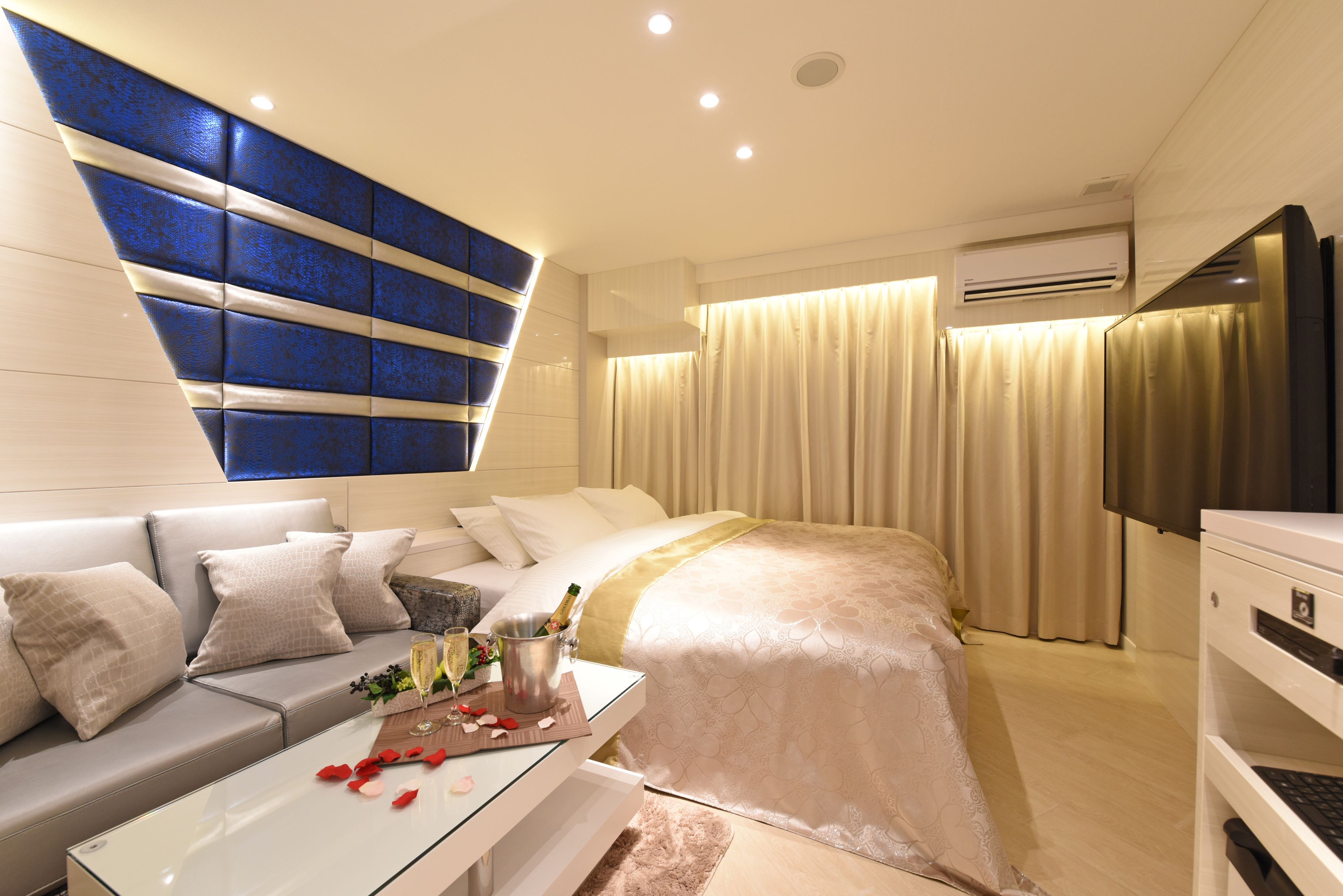 A modern hotel room with a sofa, a glass coffee table with rose petals and champagne, a bed with a gold bedspread, and a large TV.