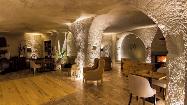 A spacious interior with high ceilings and natural stone walls, featuring a fireplace, seating areas, and a wooden floor.