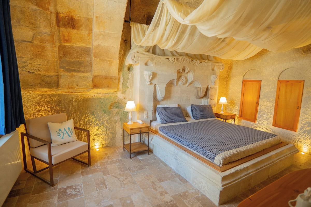 A bedroom with a stone wall, a canopy bed, a chair, bedside tables, and wooden doors.