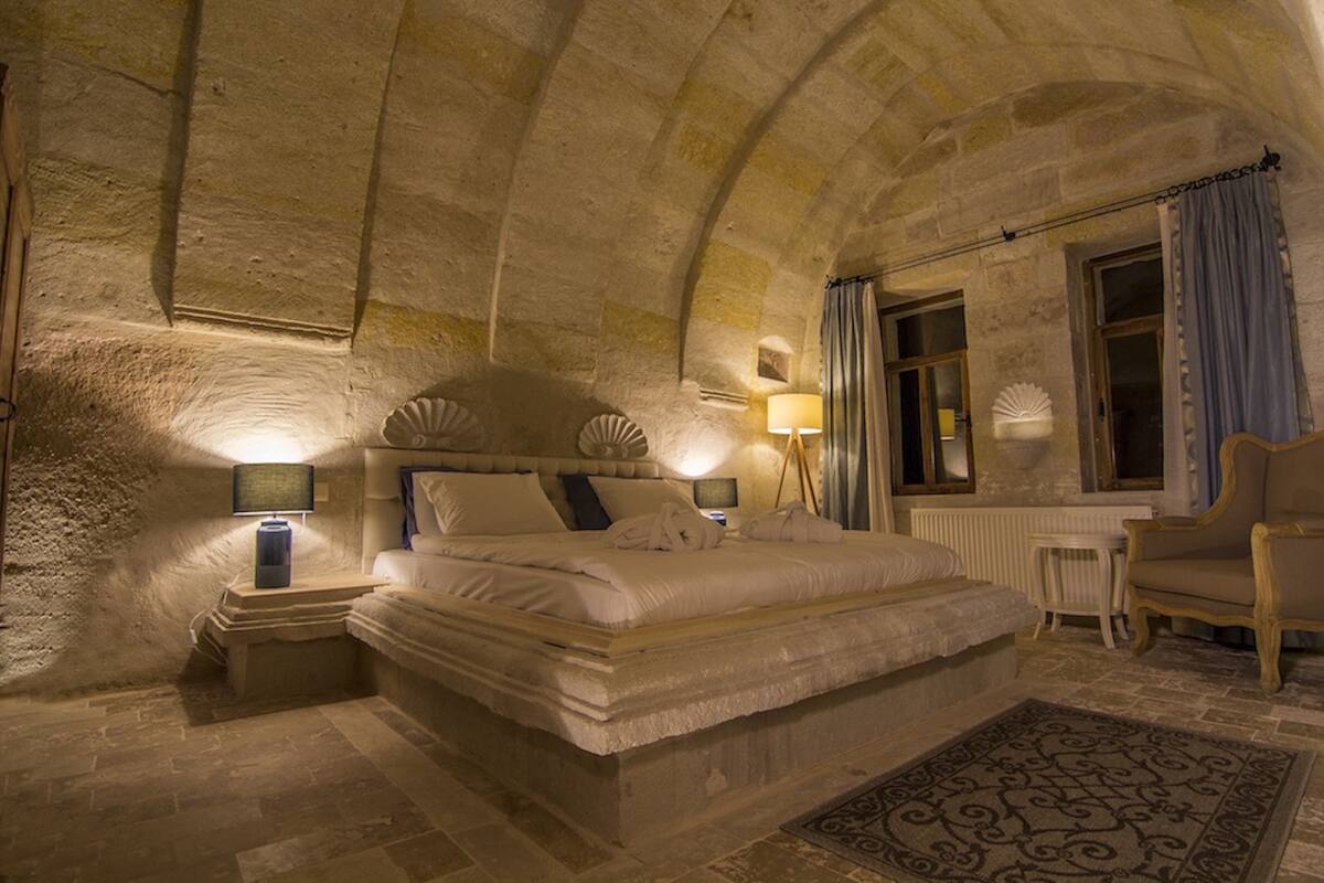 A bedroom with a stone ceiling, a large bed with pillows, a bedside table with a lamp, a chair, and a window with curtains.