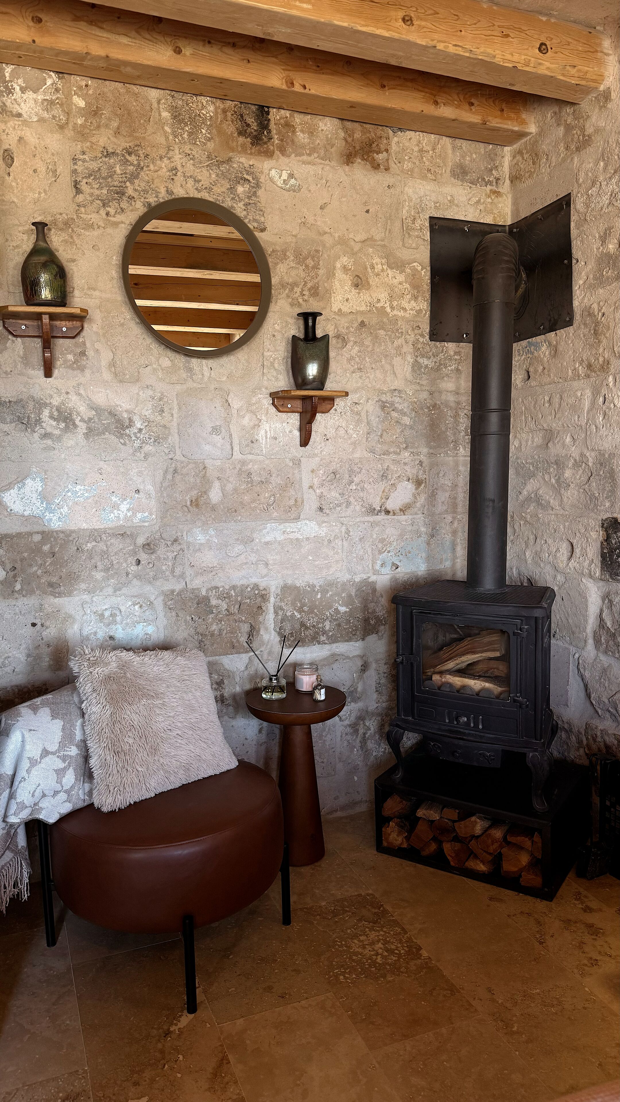 A rustic room with a stone wall, a wooden bench, a round mirror, and a black wood-burning stove.